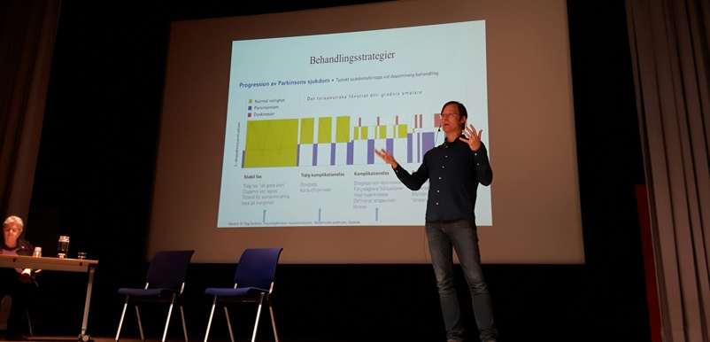 En man står på en scen och föreläser, en projektorskärm i bakgrunden