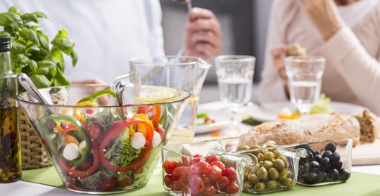 En skål med sallad i förgrunden och i bakgrunden sitter ett äldre par som äter mat