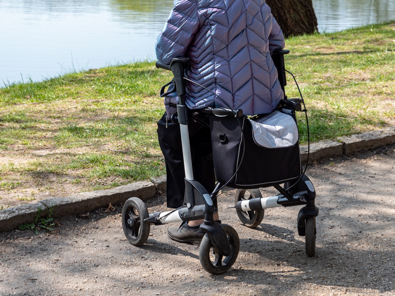En dam i täckjacka sitter på sin rollator i på en parkgång