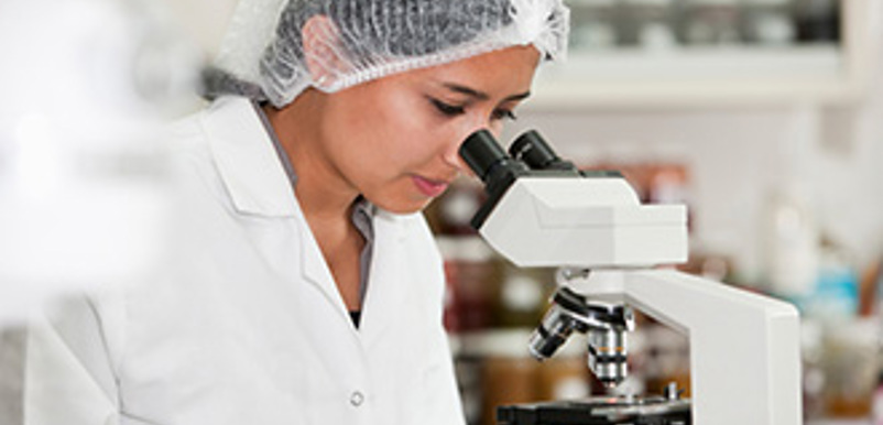 En kvinna i labbrock och hårnät tittar i ett mikroskop