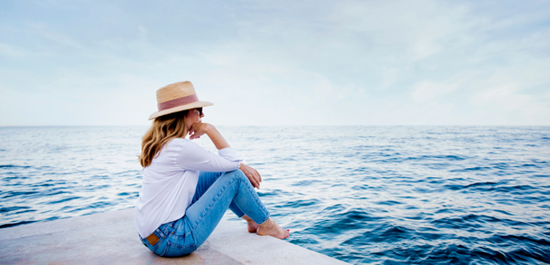 En långhårig kvinna i hatt sitter på en brygga och tittar ut över havet